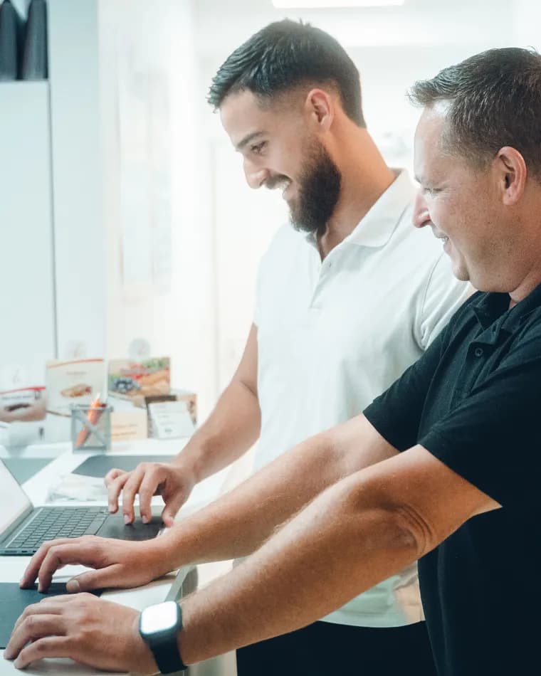 Semi Kochta und Jörg Goßler arbeiten lachend gemeinsam am Laptop in der Praxis