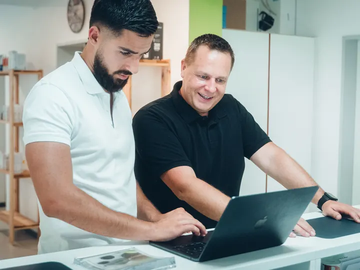 Semi Kochta und Jörg Goßler lachen gemeinsam bei der Arbeit am Laptop in der Praxis